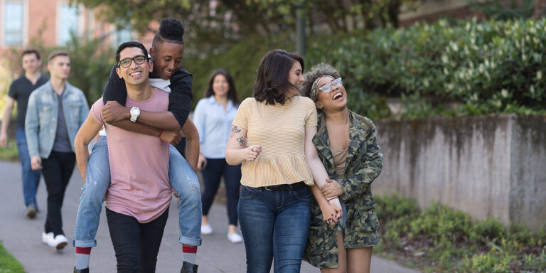 A multi-racial group of university students cheerfully walk down a sidewalk together. One student is carrying his boyfriend on his back.
