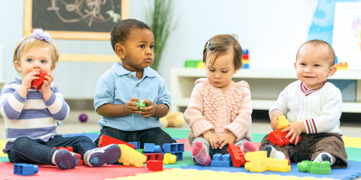 Four diverse children smiling and playing with building blocks on a colourful mat in a classroom setting.