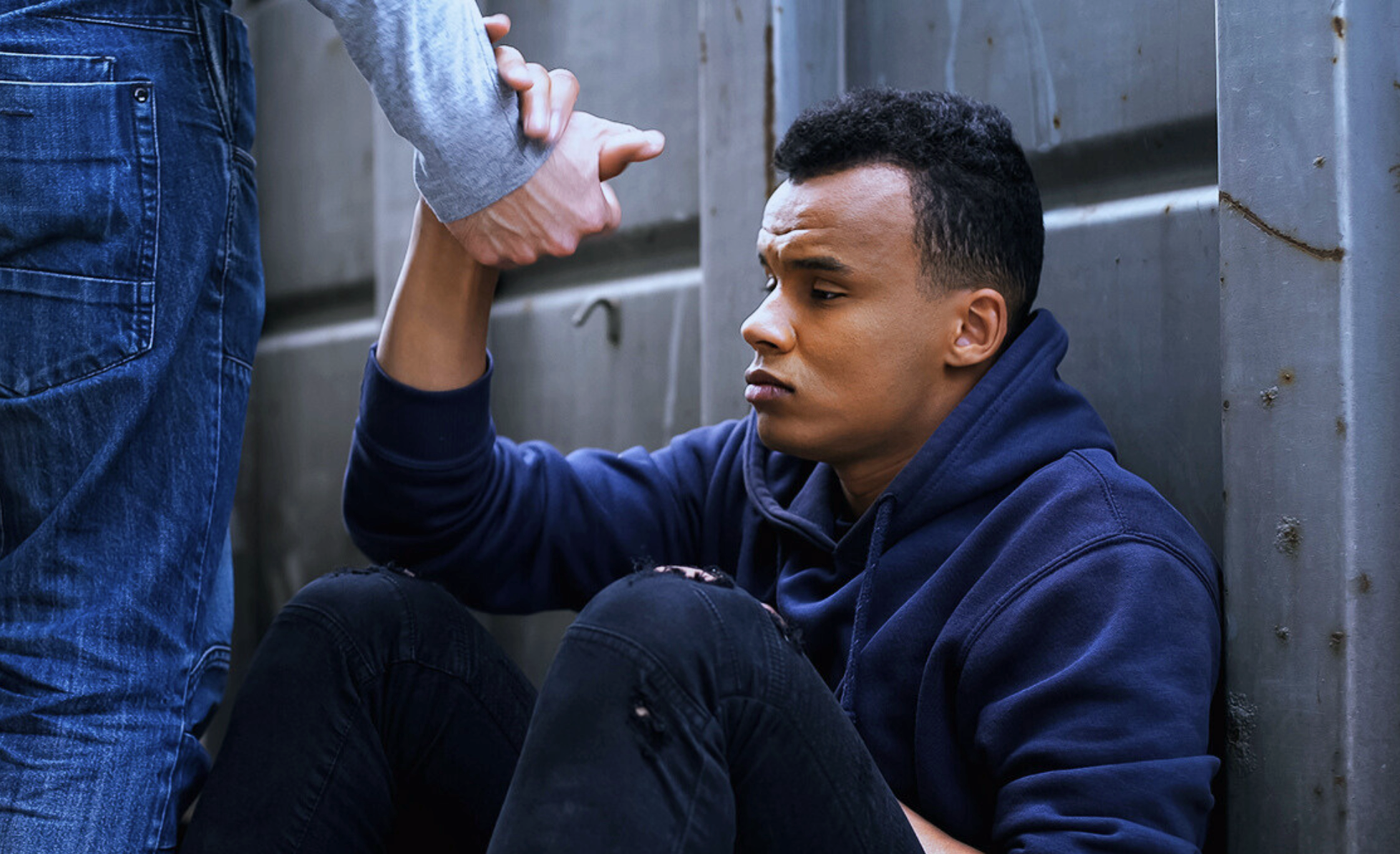 A young man sitting against a wall and receiving help from another person to stand up.