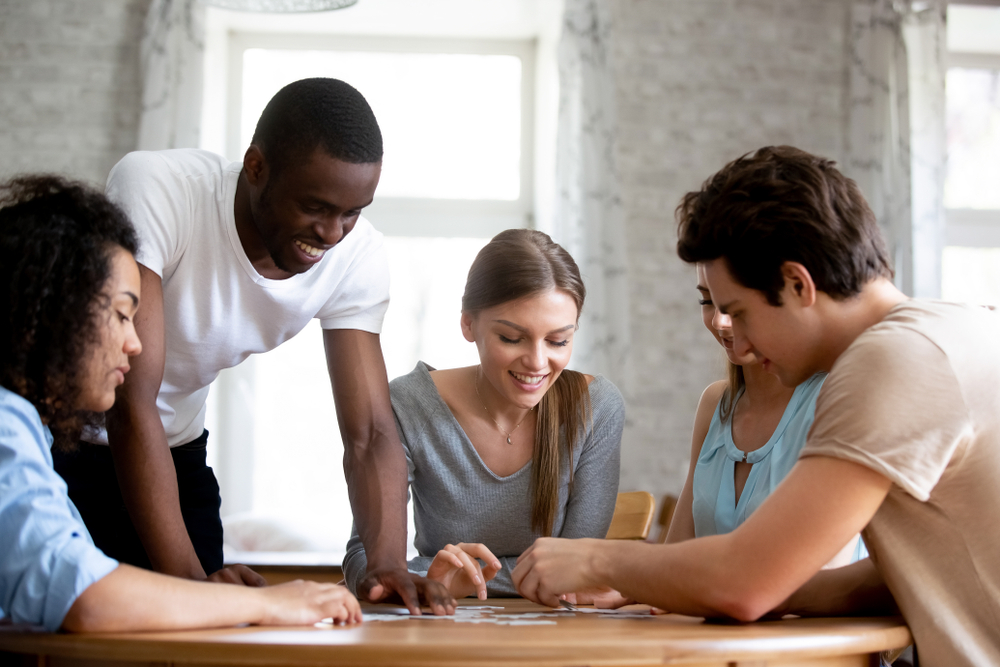 A diverse group of adults smiling while working together to assemble a puzzle at a table.