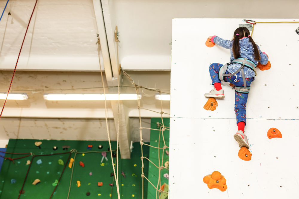 A young girl climbing an indoor rock wall while wearing a safety harness.