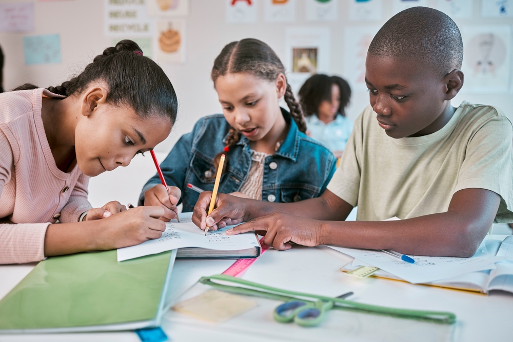 Three youth collaborating on a paper activity with school supplies on a table with a teacher sitting in the background.