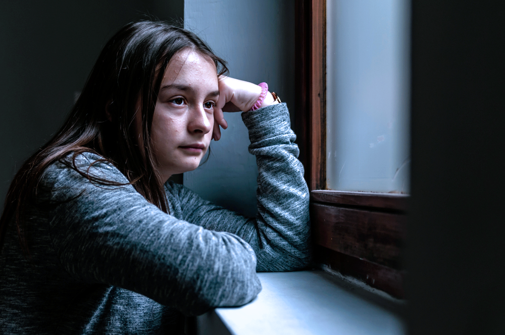 A young girl resting her hand on her head while looking out a window.