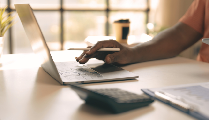 A person working on a laptop with a pen, calculator, and clipboard on a desk.