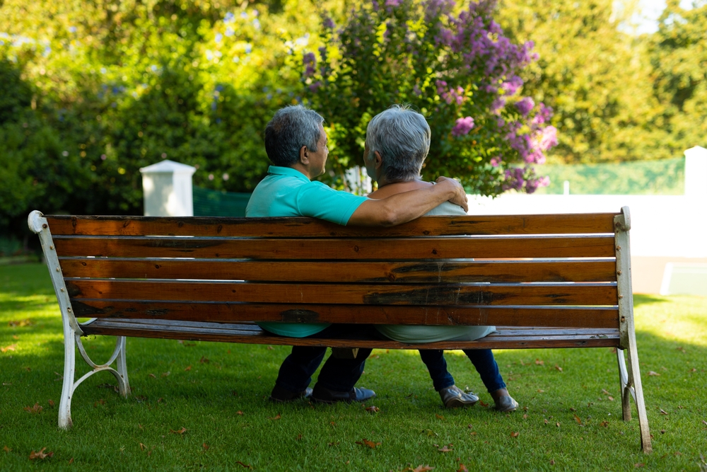 An old couple sitting on a park bench, with the man's arm around his partner as the look into the distance.