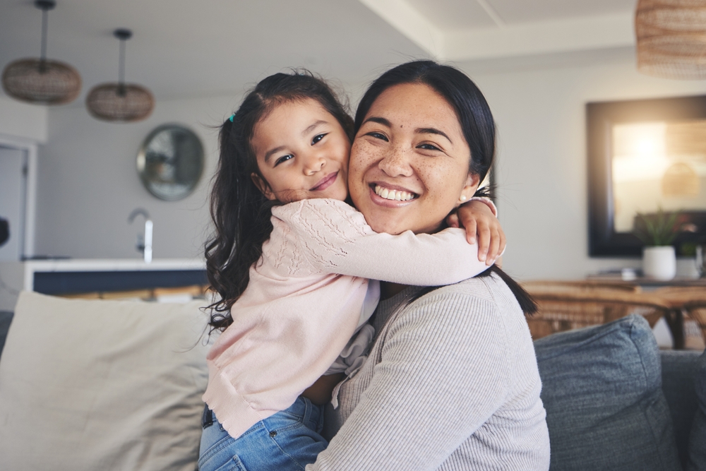 A young mother hugging her daughter while sitting on a couch, both smiling.