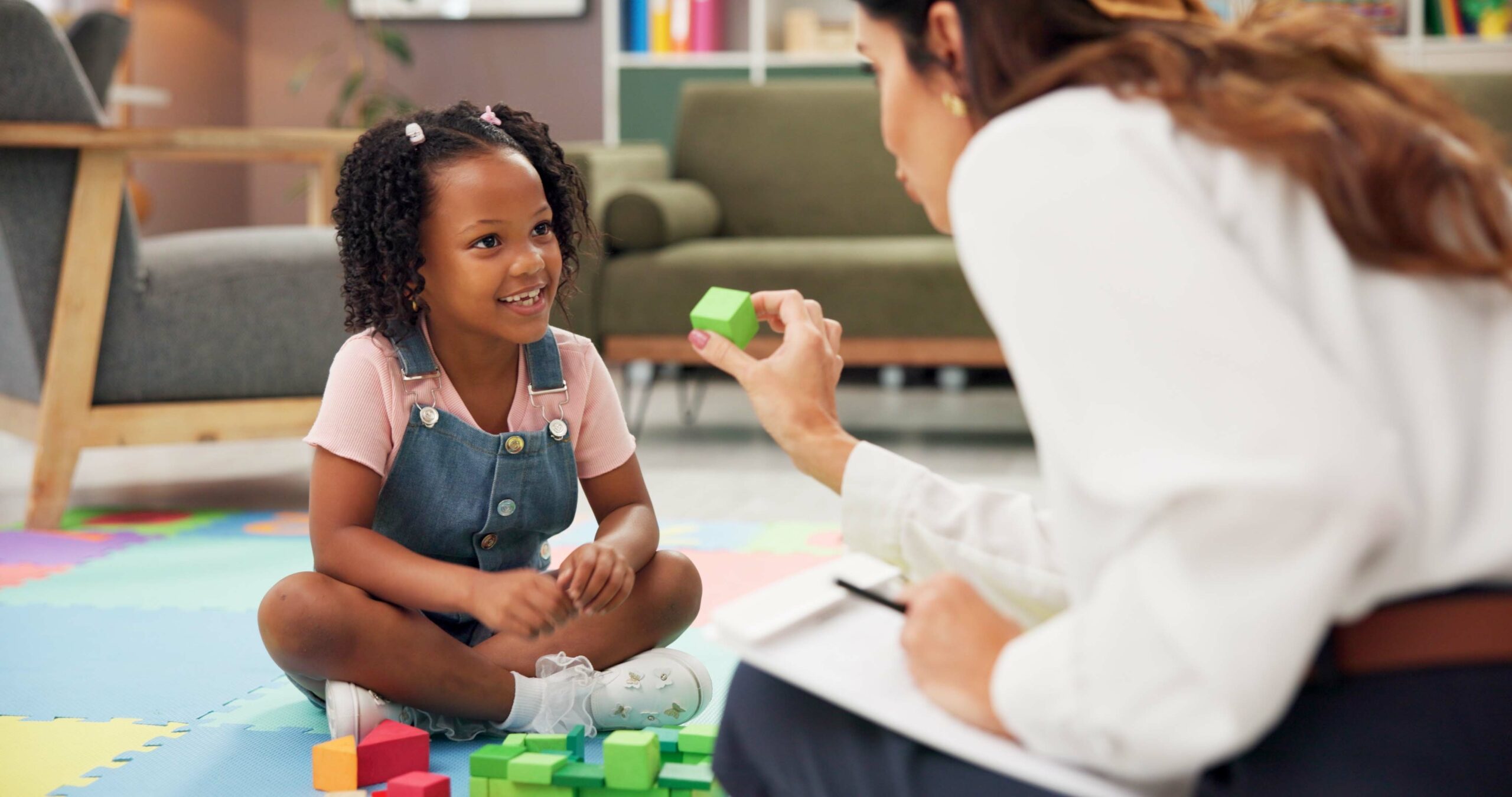 A smiling child sitting on a colourful mat with a woman showing her a square green building block.