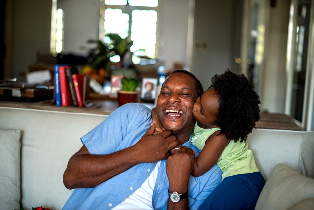 A smiling man sitting on a couch at home as a child hugs him and kisses his cheek.