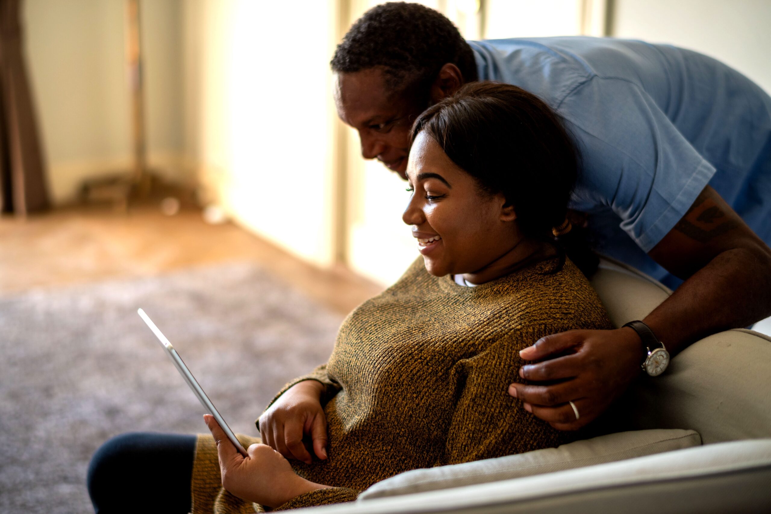 A young couple sitting on a couch at home, smiling at a tablet.